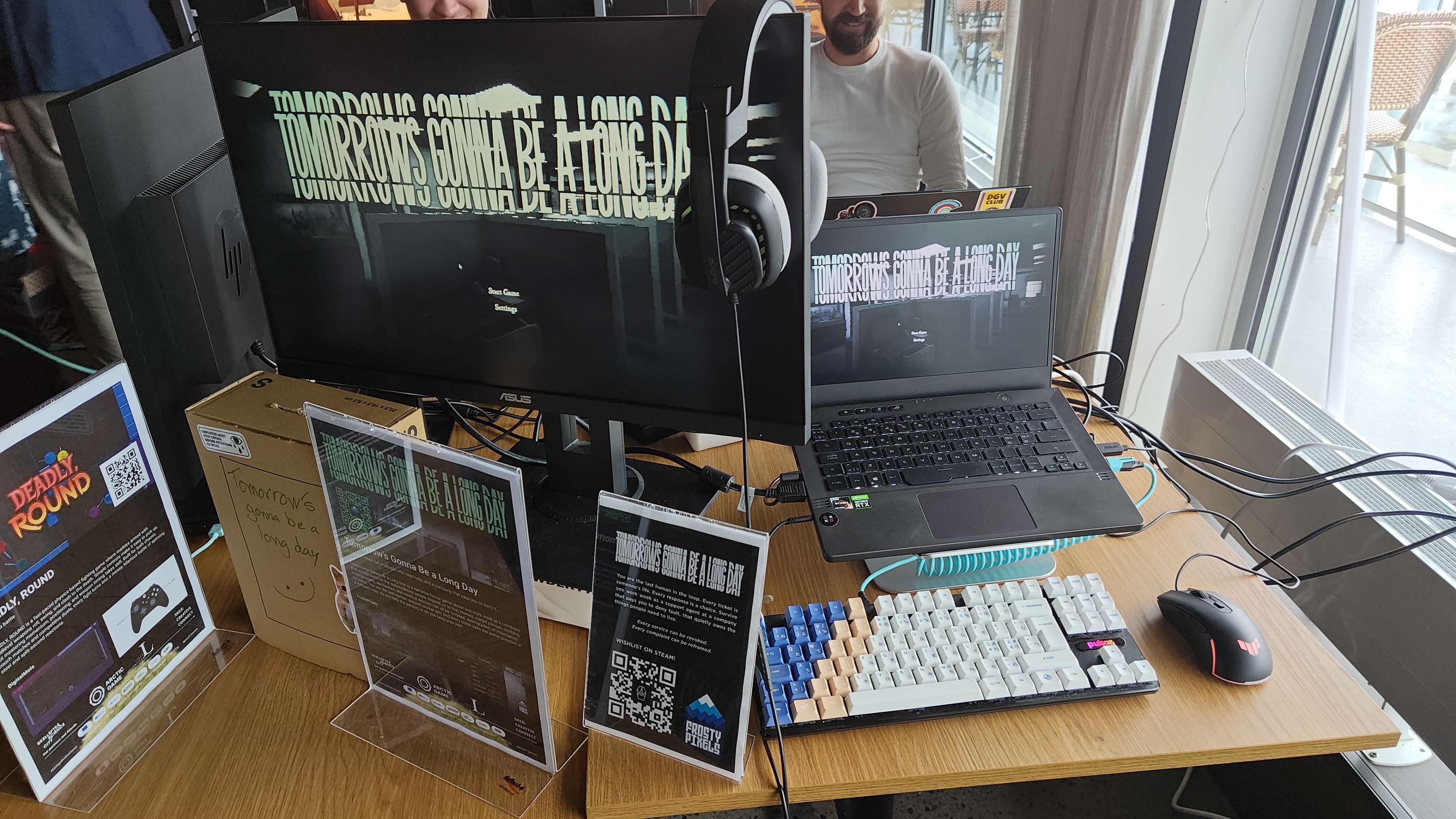 Showcase table with a monitor and a laptop both sitting on the game's main menu, headphones, keyboard and mouse, and printed standees carrying the pitch text and a Steam wishlist QR code